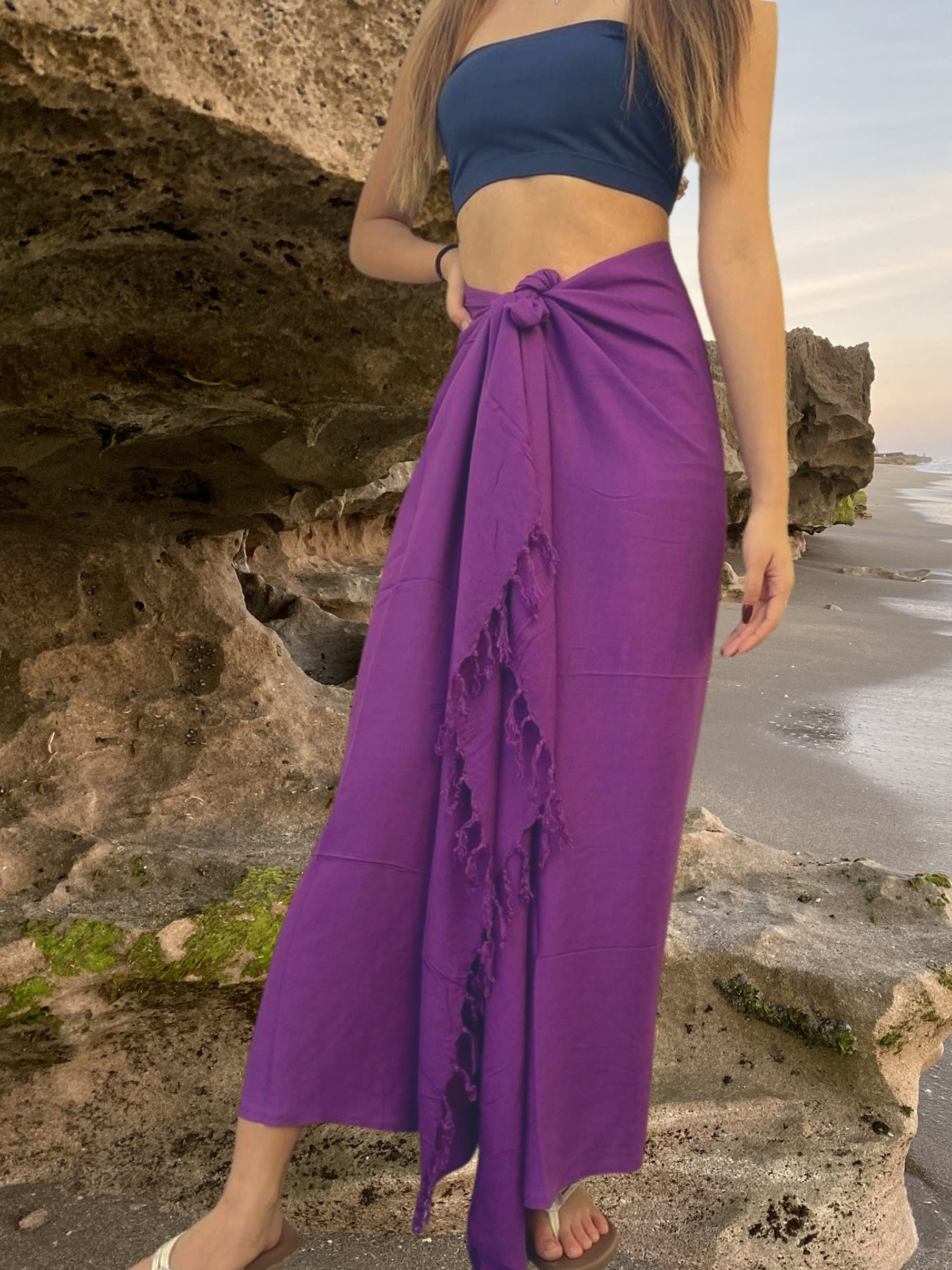 Person wearing a blue top and purple sarong standing on a rocky beach.