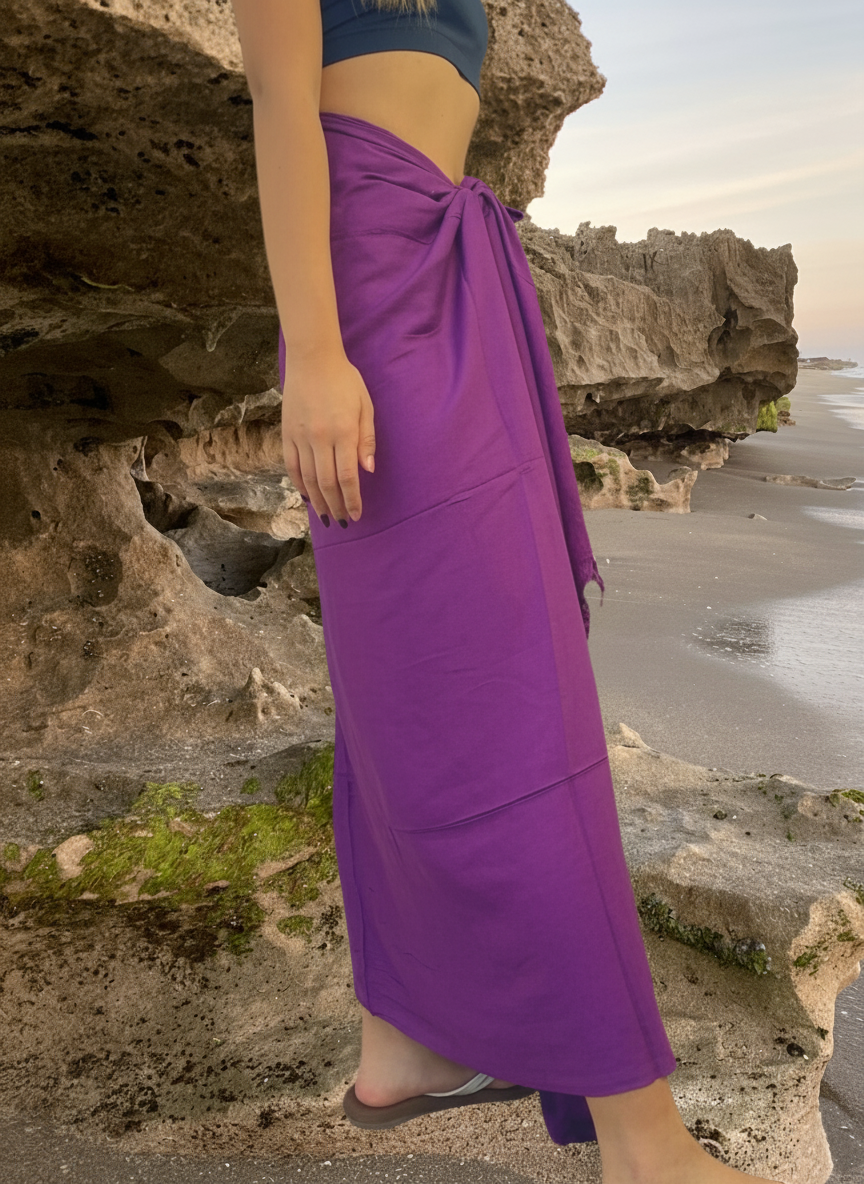 Person wearing a purple sarong standing on a rocky beach with cliffs and ocean in the background.